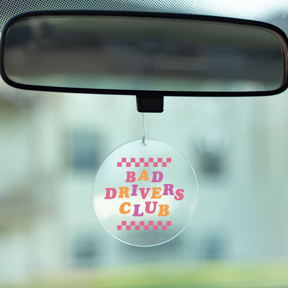 Bad Drivers Club Rearview Mirror Hang - A clear acrylic circle with the phrase &quot;Bad Drivers Club&quot; printed in pink, orange and purple text attached to a car rearview mirror with and elastic loop.