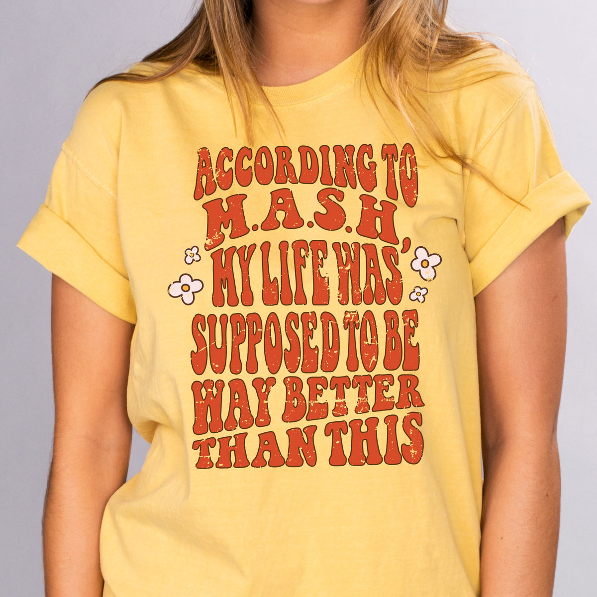 According To MASH - Femfetti - A mustard color unisex fit graphic tee with orange/red text saying &quot;According to MASH my life was was supposed to be way better than this&quot;.