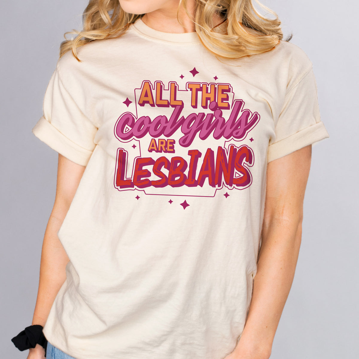 All The Cool Girls Are Lesbians - Femfetti - A natural color unisex fit graphic tee featuring the phrase &quot;all the cool girls are lesbians&quot; in the lesbian pride colors of orange purple and red.